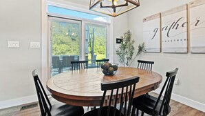 Une table de salle à manger ronde en bois, avec des chaises noires, un luminaire suspendu aux formes géométriques et un panneau mural sur lequel on peut lire : « Rassemblez-vous ».