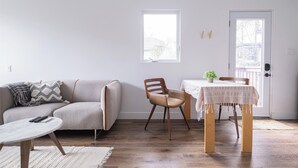 A living room with a sofa, a round coffee table, a wooden dining table with a white tablecloth, a wooden chair, and a small potted plant. - Queen size loft in nature right in the city (Toronto)
