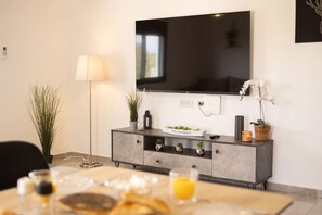 Un salon moderne avec un téléviseur à écran plat fixé au mur, un meuble gris agrémenté d’objets décoratifs et un coin repas avec une table mise pour deux personnes.