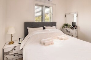 A neatly made bed with white linens and rolled towels, a marble nightstand with a lamp, and a marble dresser with a mirror and a plant.