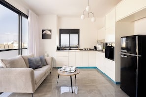 A modern living room with a sofa, a coffee table, and a kitchen area with a refrigerator and a window.
