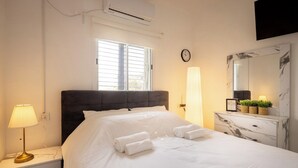 Habitación de hotel con cama, lámpara de noche, aire acondicionado, ventana, reloj y cómoda con diseño de mármol.