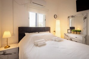 A hotel room with a bed, bedside lamp, air conditioning unit, window, clock, and a marble-patterned dresser.