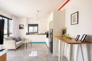 A modern living room with a sofa, coffee table, and a TV. There is a kitchen area with a fridge, sink, and cabinets.