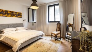 A bedroom with a bed, a chair, a mirror, and a window with curtains. - Mamad Stylish Green Oasis | Tel Aviv Old North (Tel Aviv)
