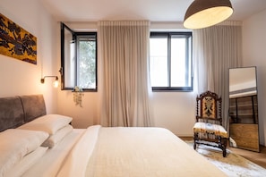 Un dormitorio con cama, cortinas, una silla, una mesita de noche y un cuadro en la pared.