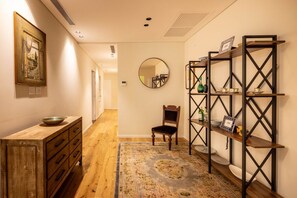Un pasillo con piso de madera, un espejo redondo, una silla de madera, un armario de madera y una estantería metálica con varios objetos.