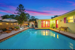 Una piscina con sillas de descanso y un pequeño solárium, rodeada por una valla de madera y una casa con fachada verde brillante y detalles en rojo.