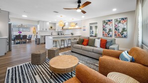 Un salon moderne avec un canapé gris, des fauteuils orange et une table basse ronde en bois. On y trouve également un coin cuisine avec des meubles blancs et un coin repas avec une table et des chaises.