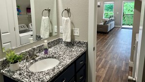 Een moderne badkamer met een granieten aanrecht, een wastafel en een spiegel. Er is een handdoekrek met twee witte handdoeken. De badkamer grenst aan een zithoek met een bank en uitzicht op groen buiten.