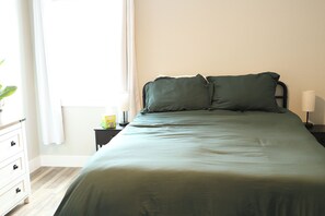 Een net opgemaakt bed met een groene sprei en twee kussens, een witte dressoir met lades, een klein nachtkastje met een lamp, en een raam met gordijnen.