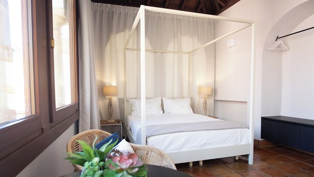A four-poster bed with white linens, a wicker chair, a small table with a plant, and a dark-colored bench. - El Alminar en Apartamentos La Casa del Aceite (Cordoba)