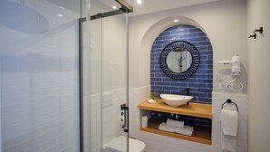 A modern bathroom with a glass-enclosed shower, a white toilet, a sink with a blue-tiled backsplash, and a wooden shelf with towels. - El Alminar en Apartamentos La Casa del Aceite (Cordoba)