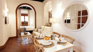 Un comedor con una mesa dispuesta para cuatro personas, rodeada de sillas de madera. Se observa una cama con dosel al fondo y una ventana redonda en la pared.