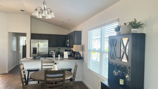 A modern kitchen with dark cabinets, a central island, and a dining area with wooden chairs. - The Sunshine Retreat in the Villages with Golf Car (The Villages)