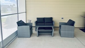 A covered patio area with wicker furniture, including a sofa and two chairs, against a light-colored wall with a window providing a view of the outside. - The Sunshine Retreat in the Villages with Golf Car (The Villages)