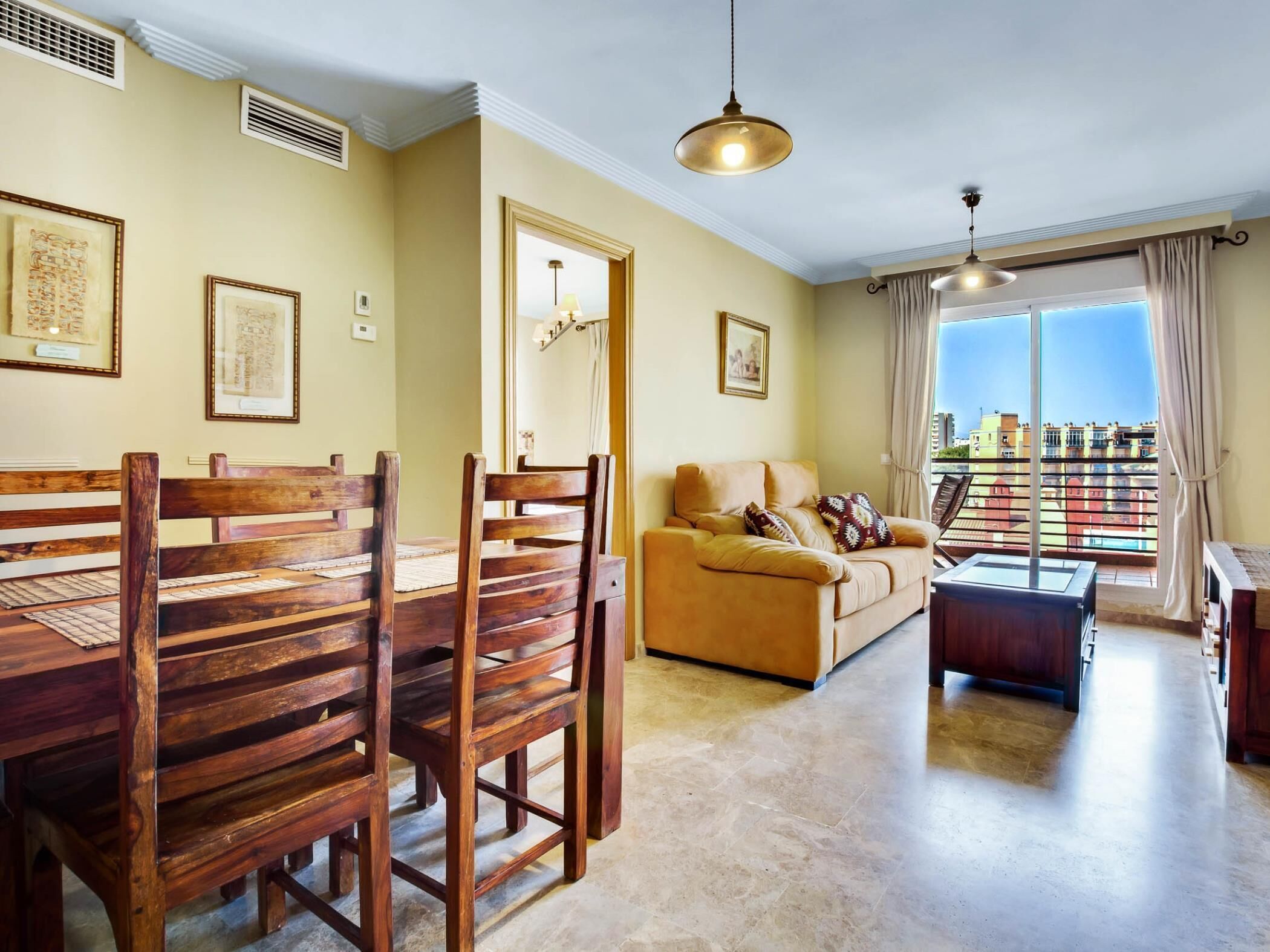 Una sala de estar con una mesa de comedor de madera y sillas, un sofá y una mesa de centro. Hay cuadros en las paredes y una vista de edificios a través de la ventana.