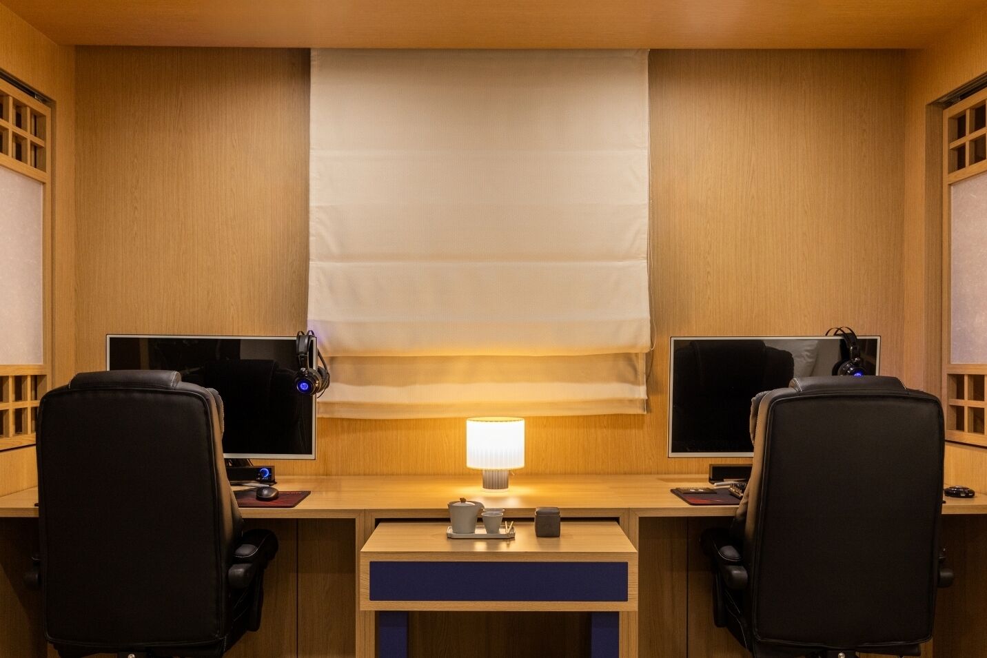 A hotel room with two workstations, each with a computer and headphones, a desk lamp, and a small table with a cup and a phone.