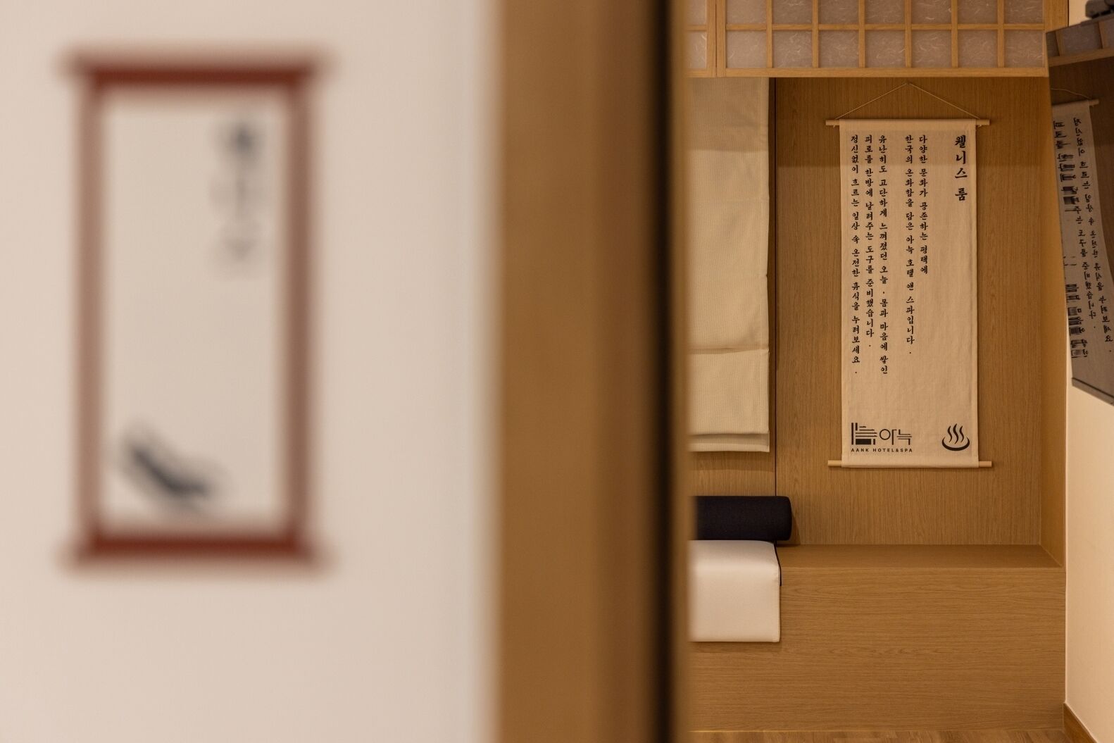 Ein Hotelzimmer mit Holzboden, einem Bett, einem Nachttisch und einem an der Wand befestigten Regal mit gerahmtem Bild und hängendem Schriftrollen.
