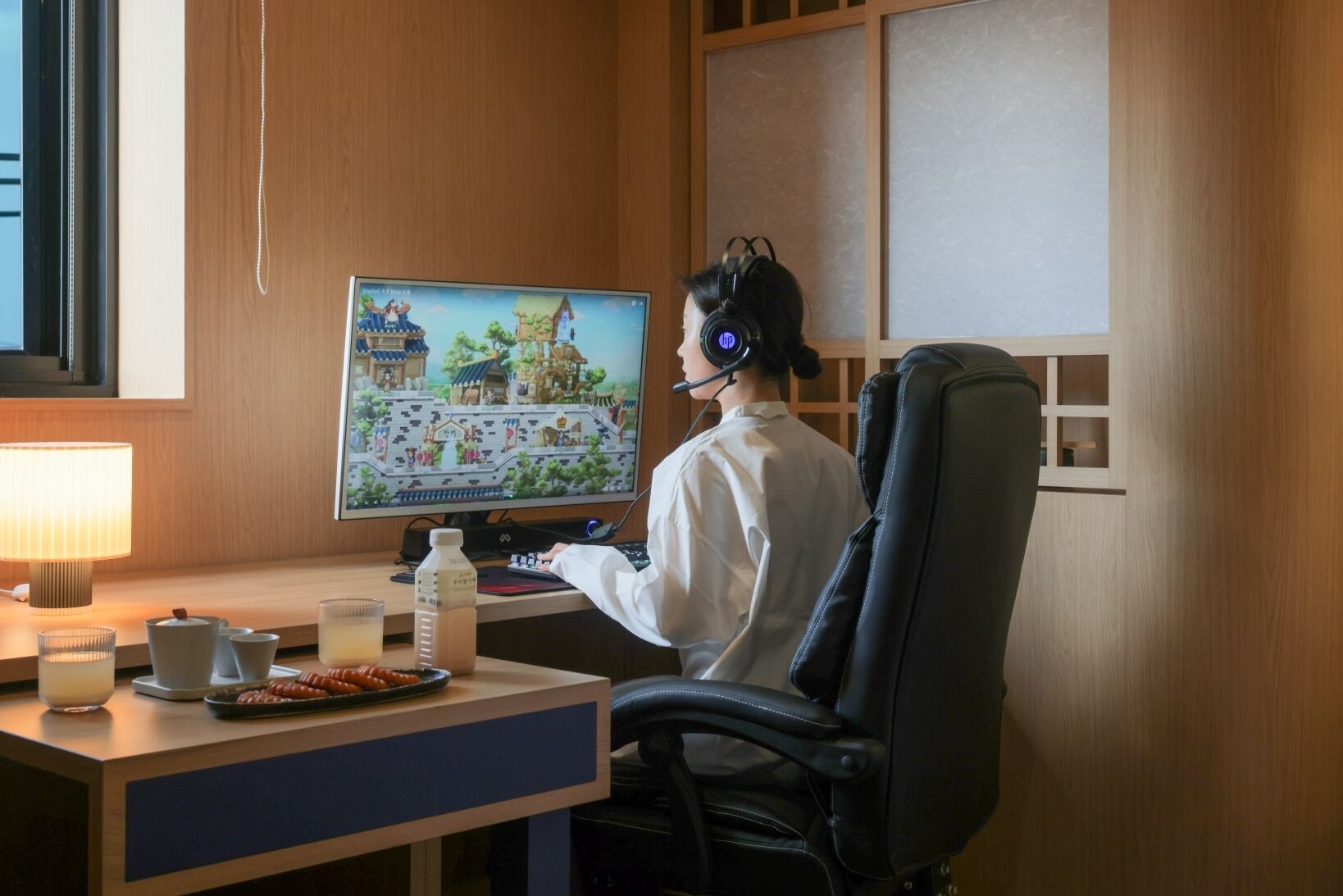 Uma pessoa sentada em uma mesa com um monitor de computador exibindo um jogo, um teclado, um mouse, um fone de ouvido e uma cadeira.
