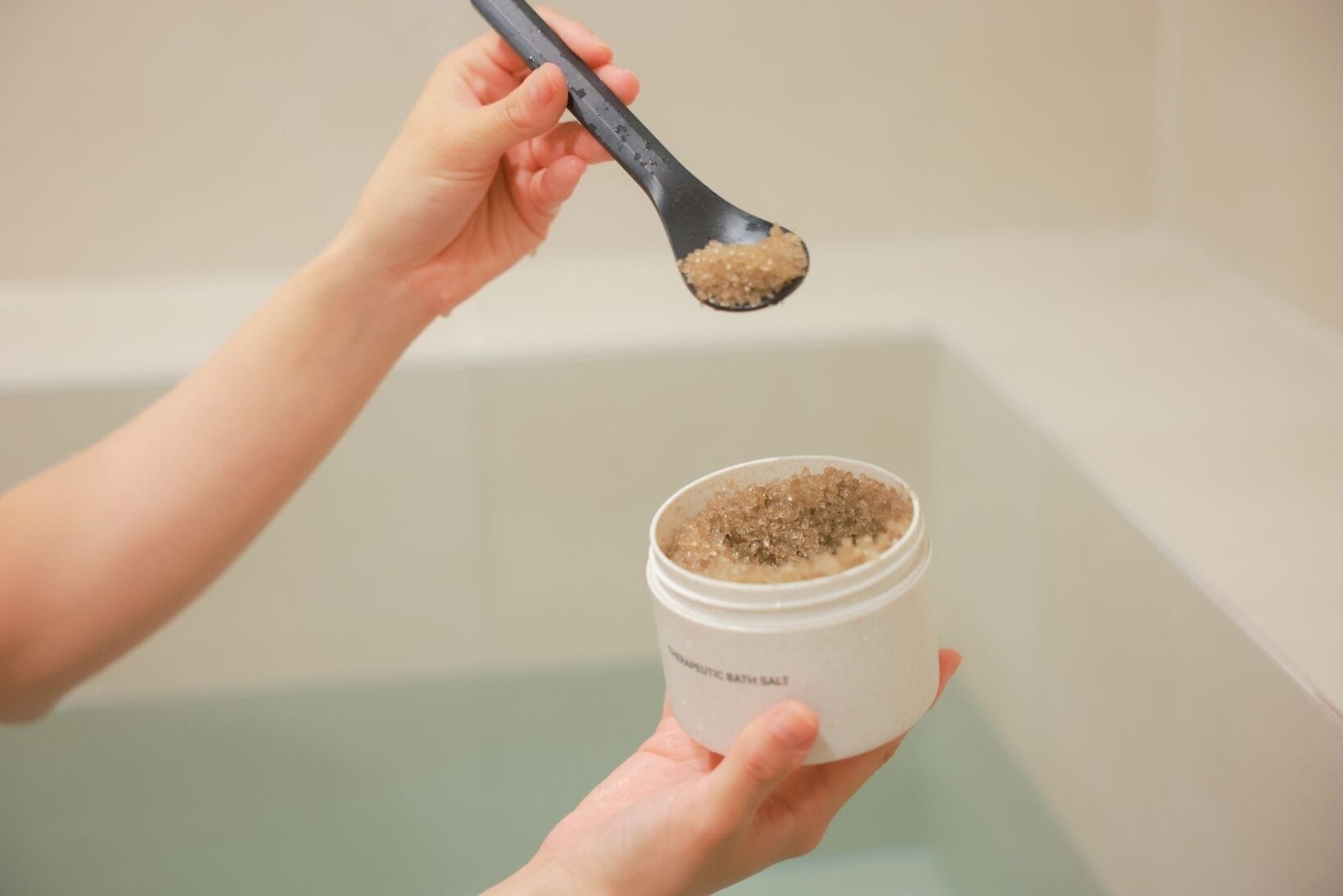 A hand holding a container labeled "BATH SALT" with a spoonful of bath salt being lifted.