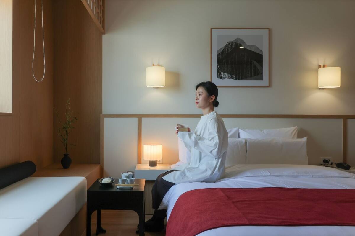 A hotel room with a bed, bedside tables, a nightstand, a wall-mounted lamp, a framed picture, and a hanging light fixture.