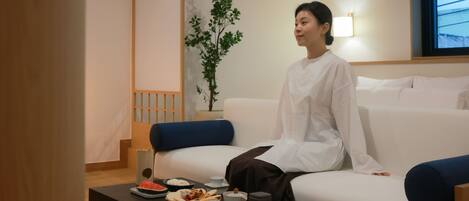 A person sitting on a white sofa in a room with a wooden floor, a small table with food and drinks, and a plant in the background.