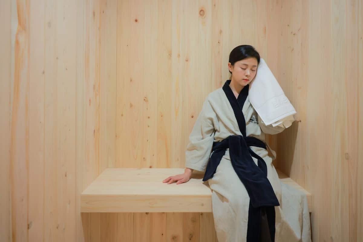A person in a traditional Japanese onsen bath wearing a white and black robe, sitting on a wooden bench with a towel over their head.