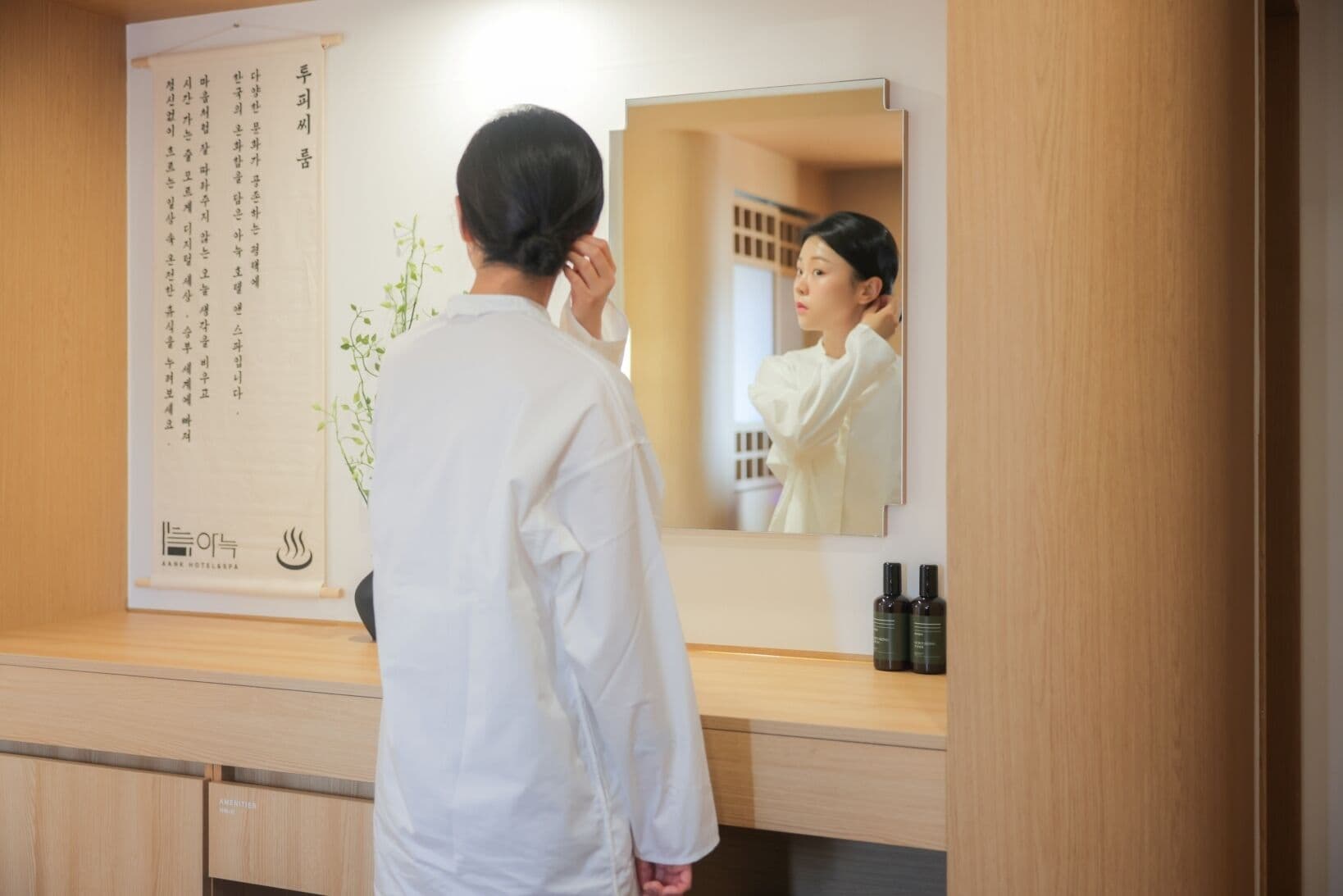 A person in a white shirt standing in front of a mirror in a room with wooden cabinets and a plant.