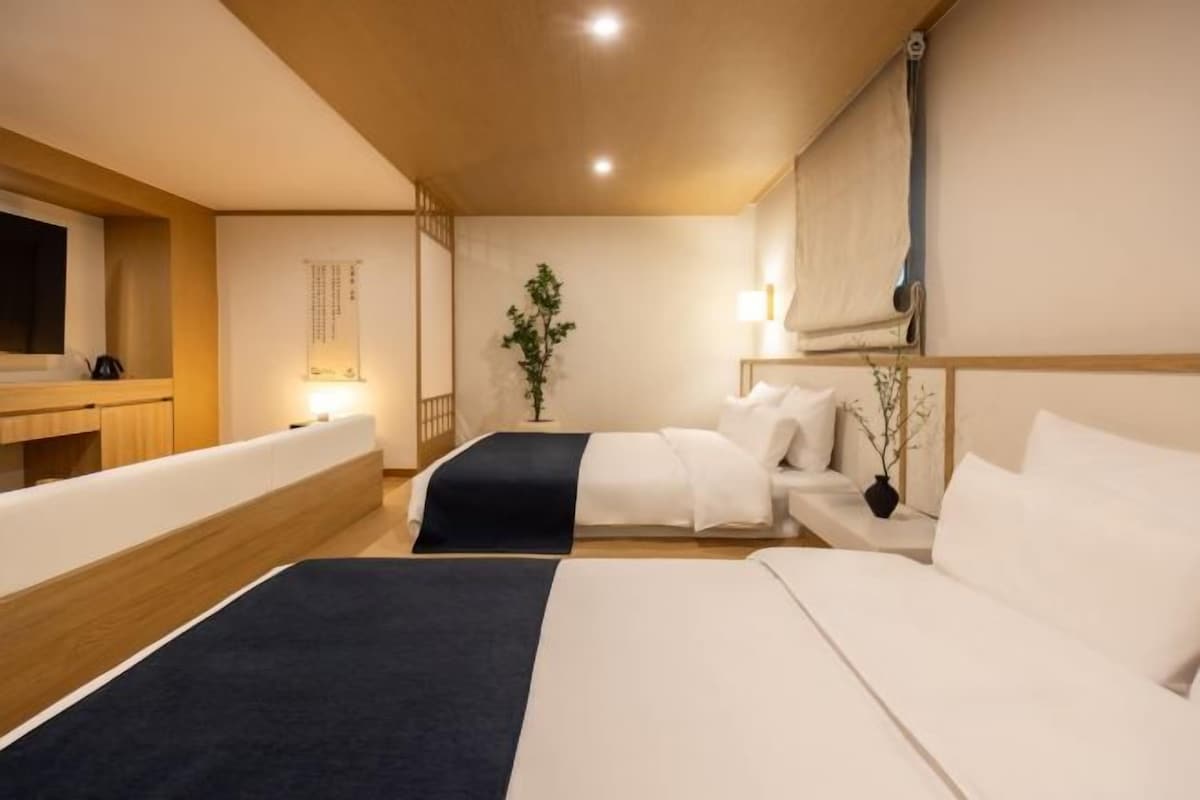 Une chambre d’hôtel avec deux lits, une tête de lit en bois, une table de chevet avec une lampe, une petite plante et un téléviseur fixé au mur.
