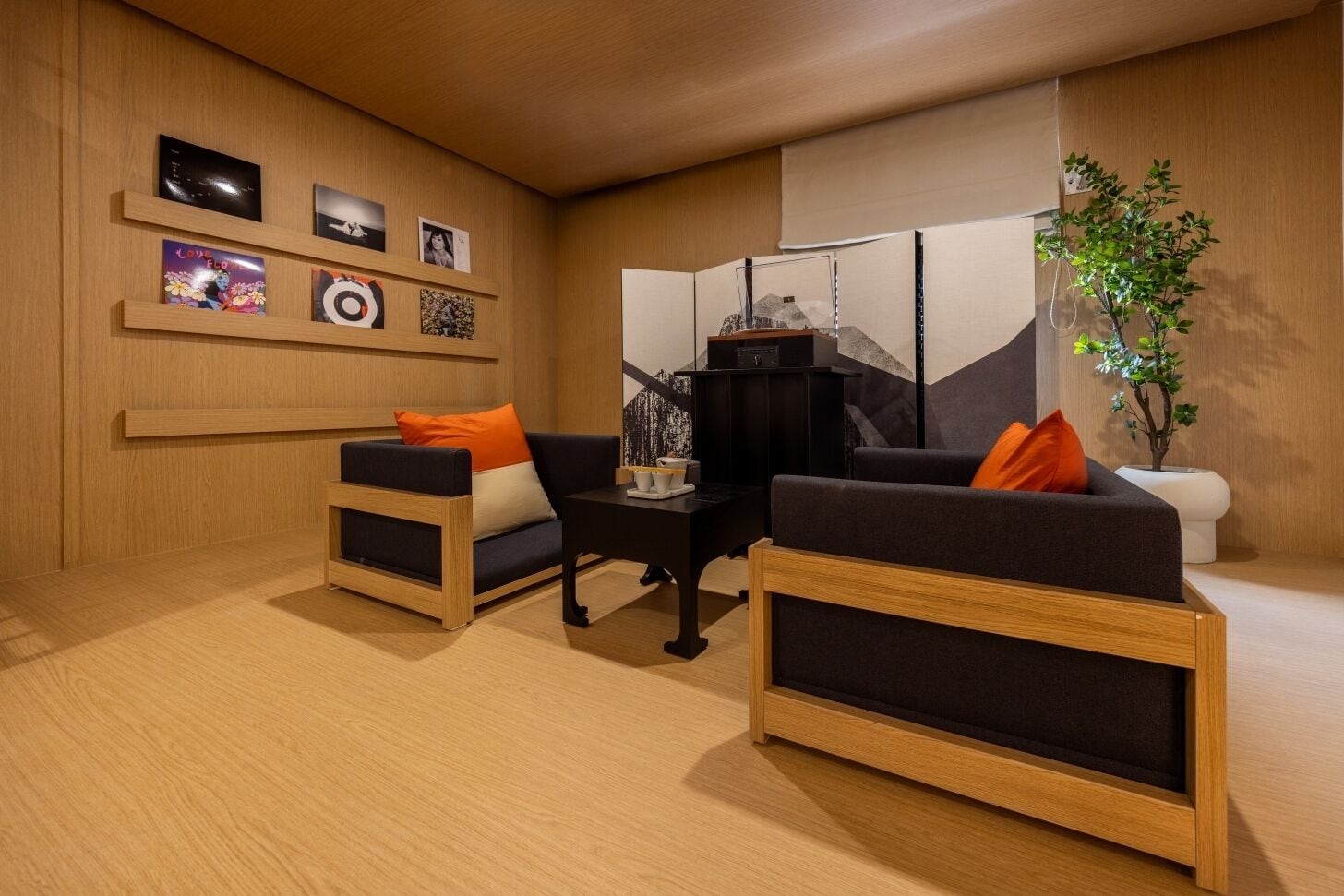Una sala de estar moderna con dos sofás, una mesa de centro y una mesita auxiliar. Hay estantes montados en la pared con cuadros enmarcados y una planta pequeña.