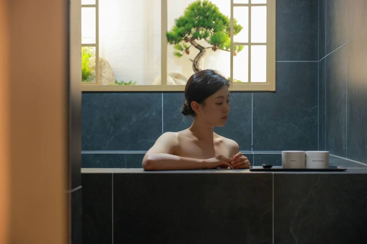 A person sitting by a window in a room with dark tiles and a bonsai tree.