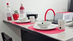A small kitchen area with a microwave, a red faucet, and a white countertop. There are two chairs and a small table with a red placemat.