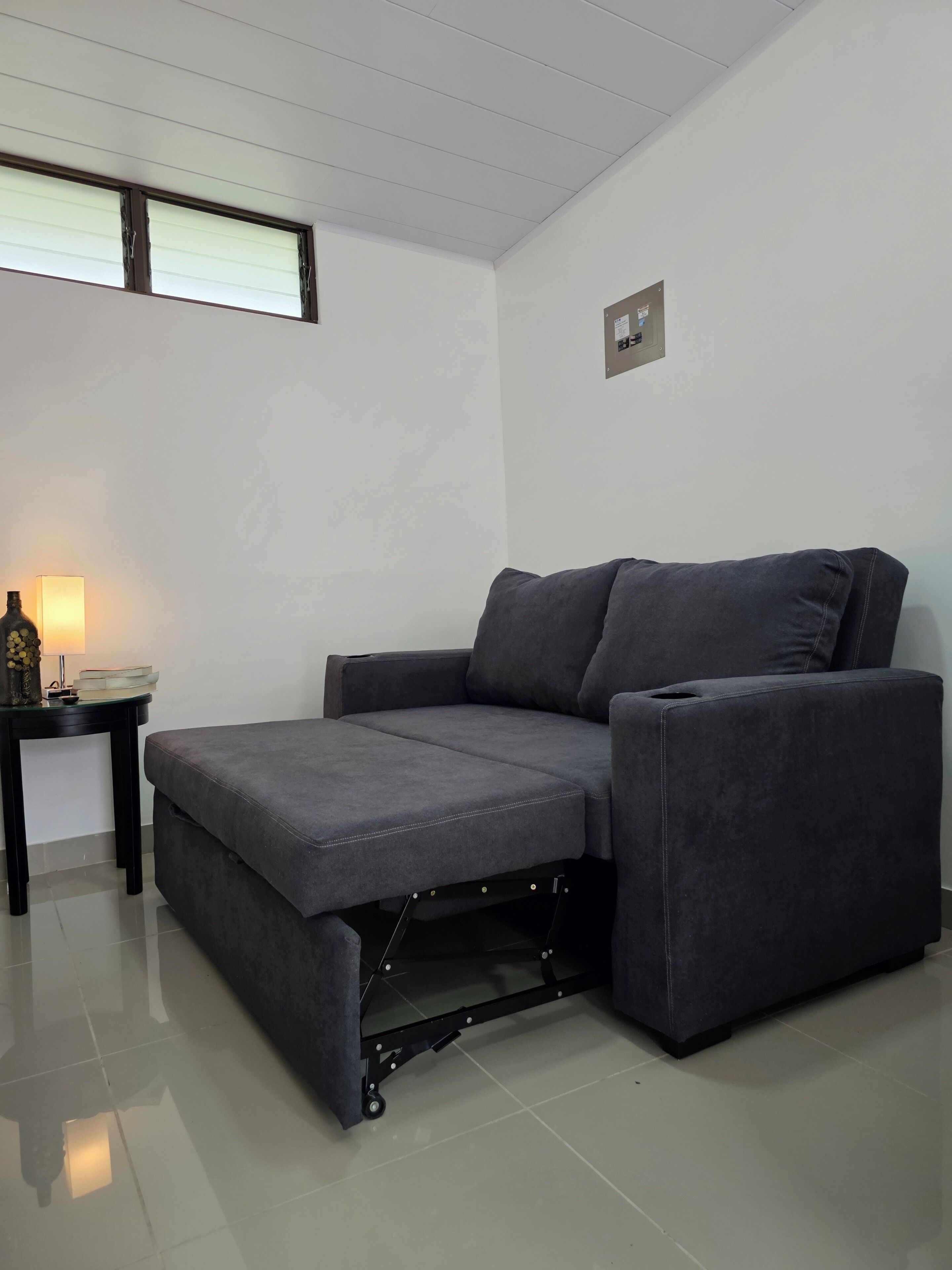 A grey sofa with a pull-out bed in a room with white walls, a small table with a lamp, and a window with blinds.
