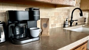 Een keuken met een Keurig koffiezetapparaat, een witte mok, een papieren zak, een spoelbak en houten keukenkastjes.