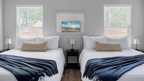 Habitación de hotel con dos camas, cada una con un edredón blanco y una manta decorativa. Hay dos mesitas de noche con lámparas y un cuadro enmarcado sobre las camas.