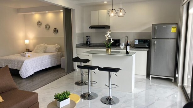 Habitació d'hotel moderna amb llit, zona de seients amb sofà i taula petita, zona de cuina amb nevera, i barra amb tamborets.