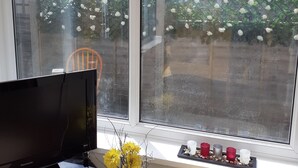 Ein Zimmer mit Fernseher, einem Fenster mit weißen Blumen, einer Vase mit gelben Blumen und einem Regal mit Kerzen.