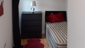Ein kleines, gut beleuchtetes Schlafzimmer mit einem Bett, einem Nachttisch mit Lampe, einem roten Teppich und einem schwarzen Vorhang.