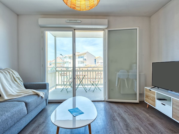 A modern living room with a grey sofa, a small white coffee table, a TV on a wooden stand, and a balcony with a view of buildings.