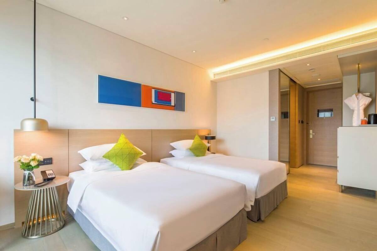 A hotel room with two beds, a bedside table with a lamp, a vase with flowers, and a modern abstract painting on the wall.