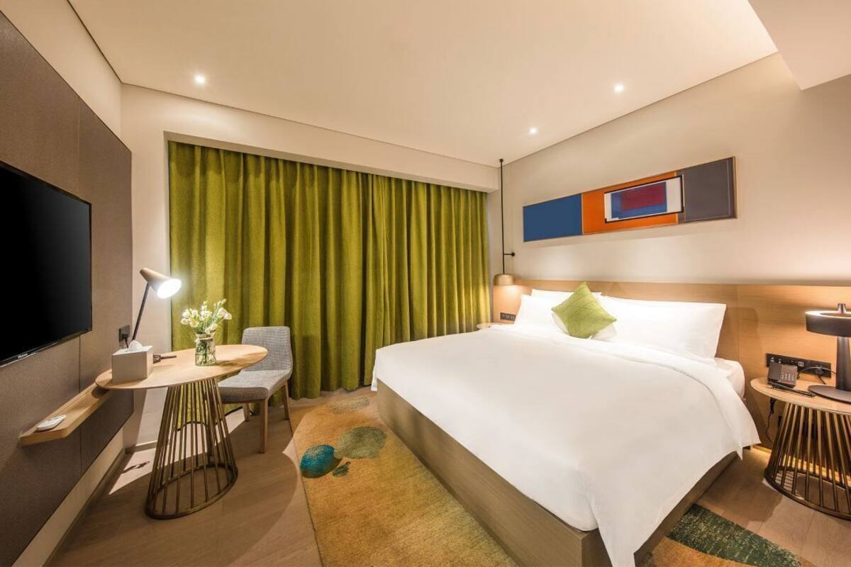 A modern hotel room with a large bed, a desk with a lamp, a TV, and a green curtain.