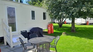 A mobile home with a patio area featuring a table and chairs, a covered grill, and propane tanks.