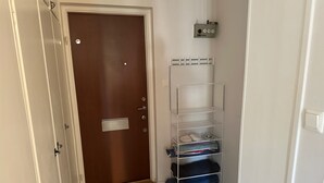 Un couloir avec une porte en bois, une étagère en métal où sont pliés des vêtements, et un thermostat fixé au mur.