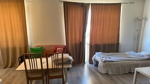 Une pièce avec un plancher en bois, un lit recouvert d’une courtepointe blanche, une table avec des chaises, un canapé avec des coussins et un lampadaire.