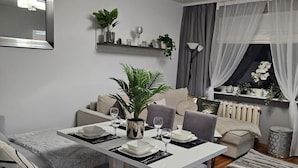 En moderne stue med spiseplads, grå sofaer, et hvidt bord og et stort vindue med lette gardiner.