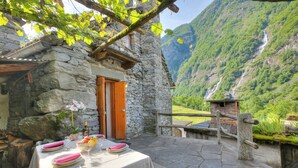 Un edificio de piedra con puerta de madera, una mesa dispuesta con platos, vasos y una botella, y vistas a una montaña y cascada.