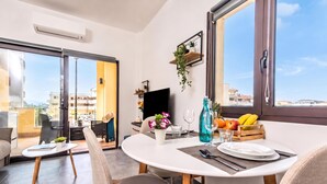 A modern dining area with a white table, cushioned chairs, a TV, and a balcony with a view.