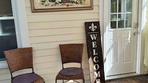 Two wicker chairs with purple cushions outside a house, a welcome sign, and a painting of flowers on the wall.