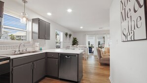 A modern kitchen with grey cabinets, a dishwasher, and a window with blinds. There is a dining area with a table and chairs, and a sofa in the background. - NEW LISTING The Proper Pelican 30A: STEPS to the BEACH! (Santa Rosa Beach)